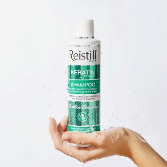 Hand holding Reistill Keratin Infusion Shampoo 250 ml with water splashes – restructuring keratin and argan oil shampoo for coarse damaged hair, hydrating lifestyle shot